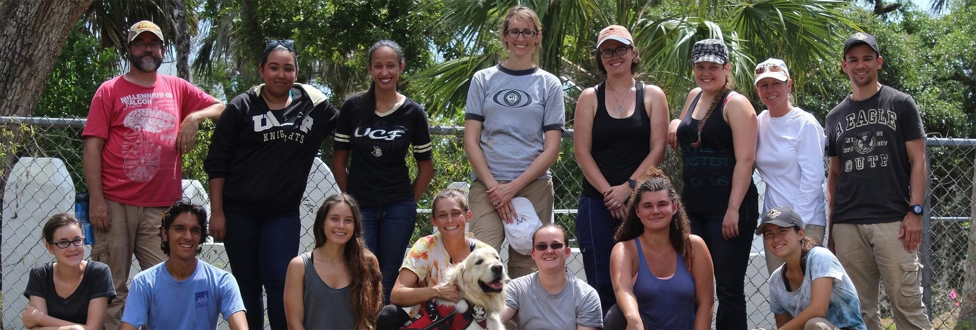 A group of 15 people, including a dog, pose outdoors in front of a chain-link fence and trees. They are smiling and dressed casually in a variety of clothing, some in hats and glasses.