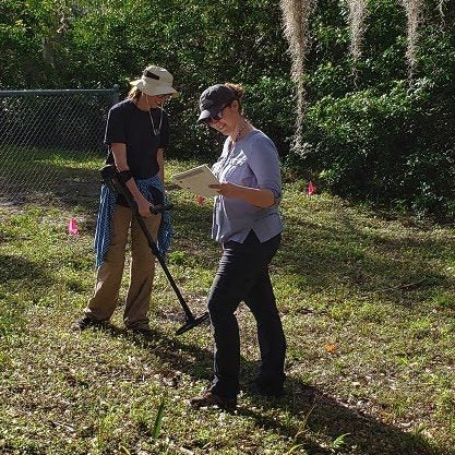 Two individuals are conducting a search in an outdoor, grassy area. One person uses a metal detector while the other reads a notepad. Both are wearing hats and casual attire. A chain-link fence is in the background.