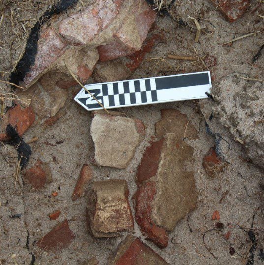 An archaeological site showing a white and black measuring scale placed among bricks and stones partially covered by sand.