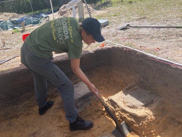 A person wearing a green "Florida State Parks" shirt and black cap is using a shovel to excavate a square pit in a sandy area. Tools and equipment are visible in the background.