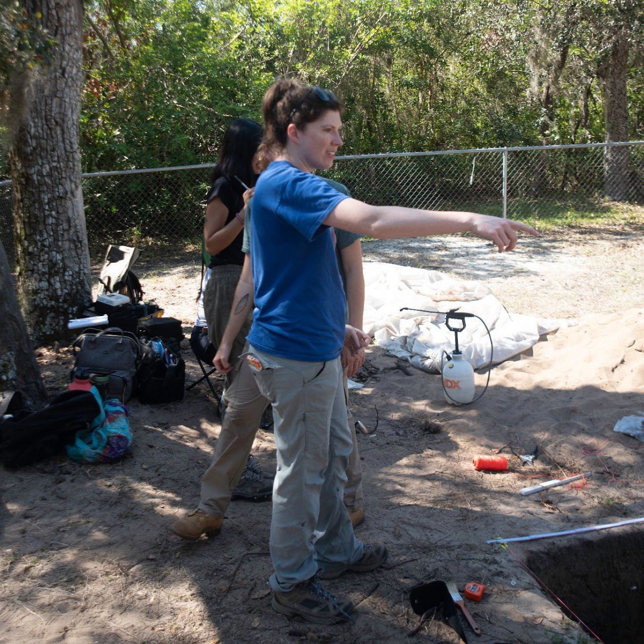 Two people stand near an excavation site with one person in a blue shirt pointing. The area is surrounded by trees and tools are scattered on the ground.