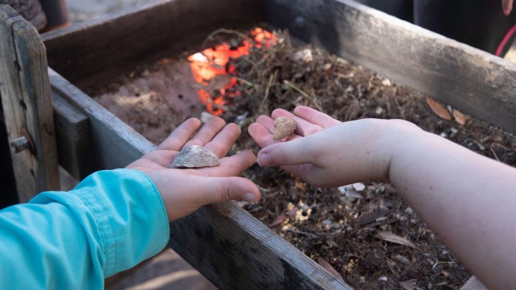 Two hands hold shells near a wooden sifting tray filled with dirt and debris, with a red glow appearing through gaps in the tray.