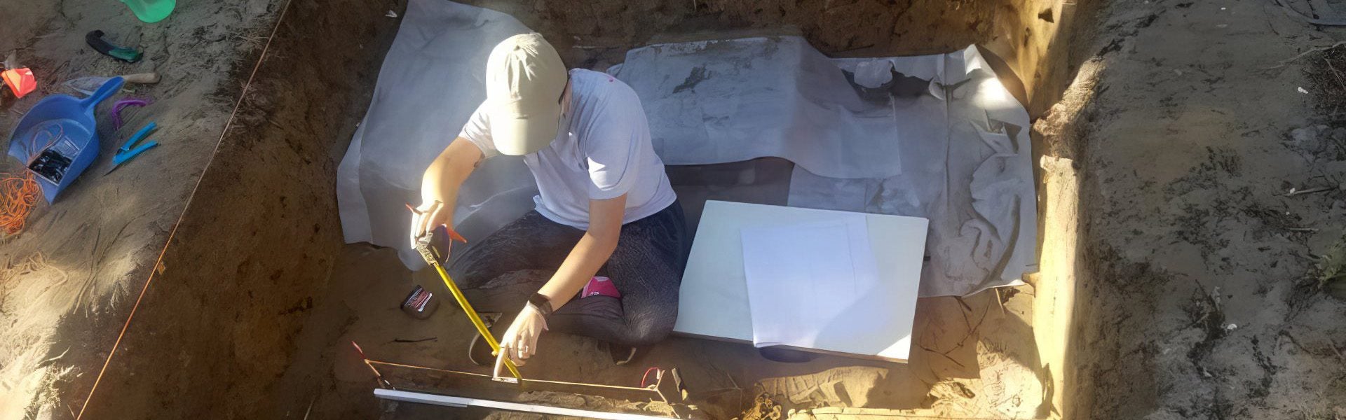 A person wearing a cap is sitting in an excavation pit, using a measuring tape, with various tools and papers around.