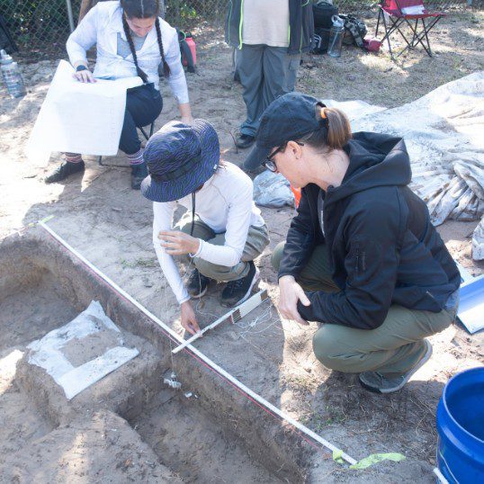 People are engaged in an archaeological excavation, measuring a grid marked area at a dig site. Two individuals are using measuring tools, while others are in the background with paperwork.