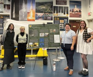 Four individuals stand in front of a display board featuring detailed information and images about space exploration, set in an exhibition room decorated with space-themed posters and models.