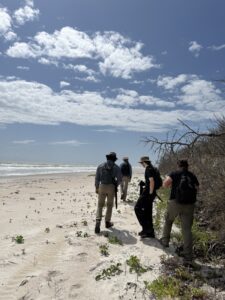 Four people wearing backpacks walk along a sandy beach under a partly cloudy sky, with the ocean on their left and sparse vegetation on their right.
