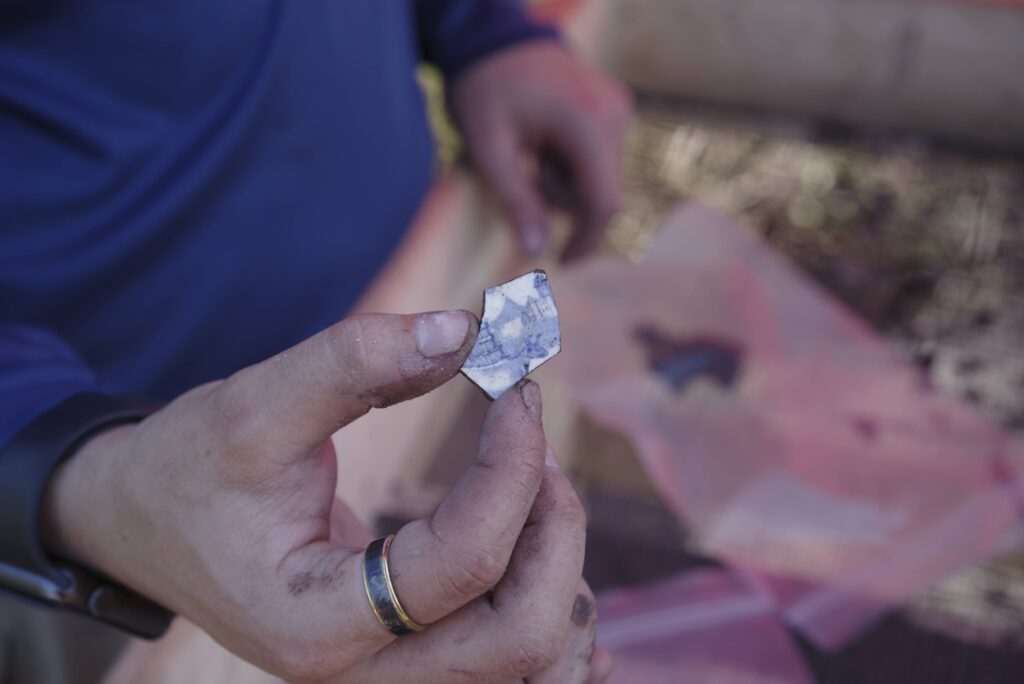 A person with dirt-stained hands holds a small, irregularly shaped, reflective object, possibly a mineral or artifact, over a table with plastic bags and other items.