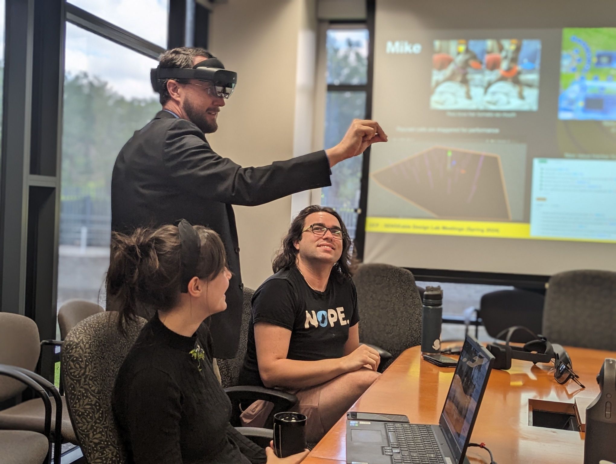 A man is using augmented reality glasses while pointing at something in the air during a presentation. Two other individuals are seated, watching, with a laptop and a projector screen displaying slides.