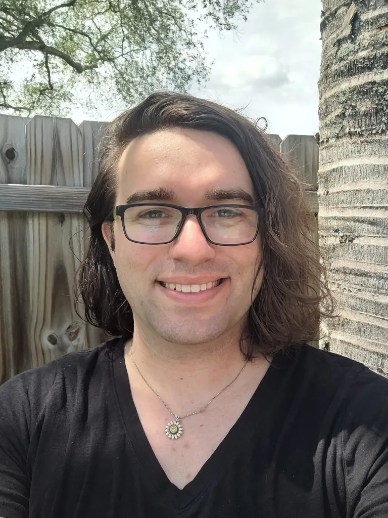 A person with long hair and glasses smiles at the camera, standing outdoors near a wooden fence and a palm tree.