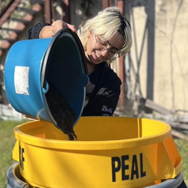 Woman pouring liquid from a blue bucket into a large yellow and gray industrial container, with stairs and greenery in the background.