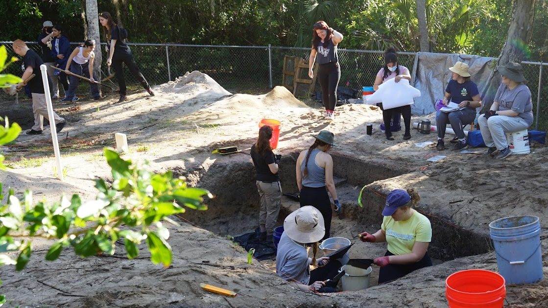 Group of people, some wearing hats, participating in an outdoor archaeological dig, with tools and buckets, in a wooded area.