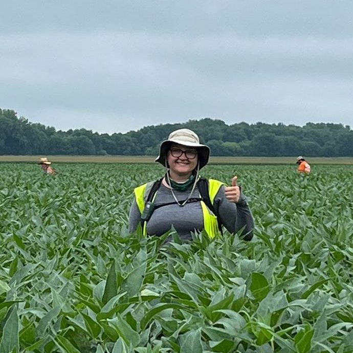 Person in a hat and backpack giving a thumbs up while standing in a lush green soybean field, with two other people visible in the background.