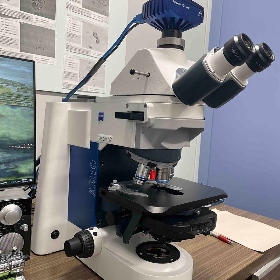 A high-resolution microscope on a desk, equipped with multiple lenses and adjustment knobs, next to a computer mouse and scientific posters on the wall.