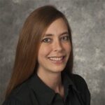 A person with long brown hair, wearing a black shirt, smiles at the camera against a gray background.