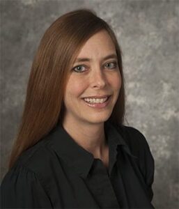 A person with long brown hair, wearing a black shirt, smiles at the camera against a gray background.