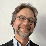 A man with glasses, gray hair, and a beard smiles at the camera, wearing a gray shirt and dark vest, against a plain background.