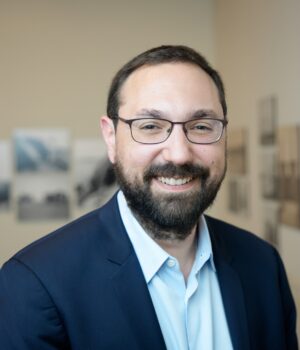 A man with glasses and a beard, wearing a dark jacket and light shirt, stands smiling in a gallery with photographs on the wall behind him.