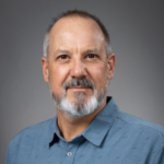 A middle-aged man with short hair, a gray beard, and mustache, wearing a blue button-up shirt, poses against a plain gray background.