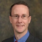 A man with short brown hair, glasses, and wearing a dark suit, blue shirt, and red tie, poses in front of a neutral brown background.