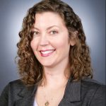 A woman with curly hair smiling in front of a gray background.