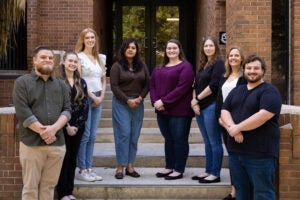 Eight adults stand in two rows on steps outside a brick building, posing for a group photo.