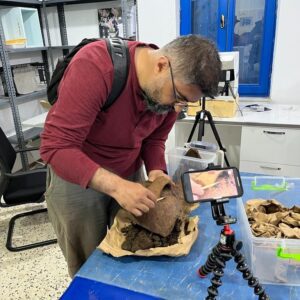 A person examines an archaeological artifact at a table while being recorded by a smartphone on a tripod.