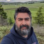 A person with a beard is wearing a dark hoodie and standing outdoors with a background of green fields and a cloudy sky.