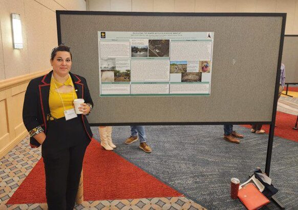 Person stands beside a research poster in a conference room, holding a cup. The poster includes images and text on a large board. Carpeted floor and other attendees in the background.