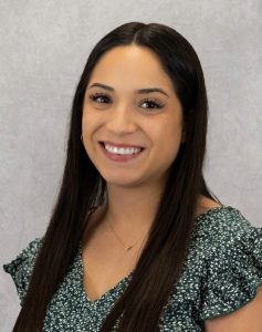 A woman with long, straight black hair wearing a green patterned blouse smiles in front of a grey textured background.