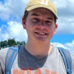 Person wearing a cap and backpack in front of an ancient stone structure with a cloudy sky.