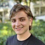 A young person with short hair is smiling while standing in front of a leafy background. They are wearing a black shirt.