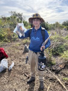 A person wearing a hat and blue shirt holds up a sealed plastic bag with an object inside. They stand in a field holding a water bottle and a notebook under a partly cloudy sky.