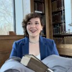 Person in a blue blazer smiles while sitting in a library, holding an open book.