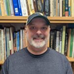 A man wearing a dark cap and gray shirt smiles in front of a shelf filled with books on various subjects.