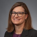 A person with shoulder-length brown hair, wearing glasses, a dark blazer, and a purple top, smiles against a plain gray background.