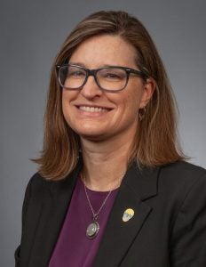 A person with shoulder-length brown hair, wearing glasses, a dark blazer, and a purple top, smiles against a plain gray background.