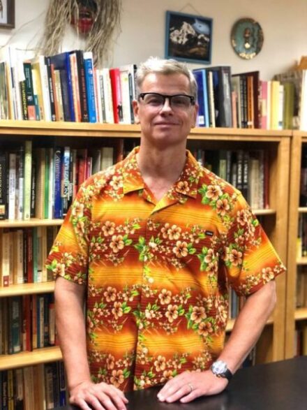 A man wearing glasses and a bright orange floral shirt stands in front of a bookshelf filled with books.