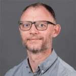 A man with glasses and a beard, wearing a gray button-up shirt, poses in front of a gray background.