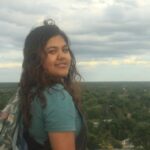 Person with long hair and a backpack stands on a hill overlooking a town, with a cloudy sky in the background.