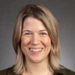 A woman with straight shoulder-length blonde hair smiles at the camera against a plain gray background.