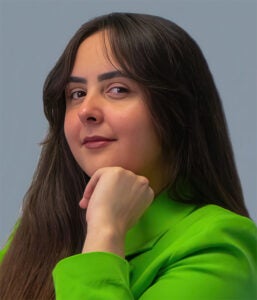 Woman with long dark hair wearing a bright green jacket poses with her chin resting on her hand against a plain gray background.