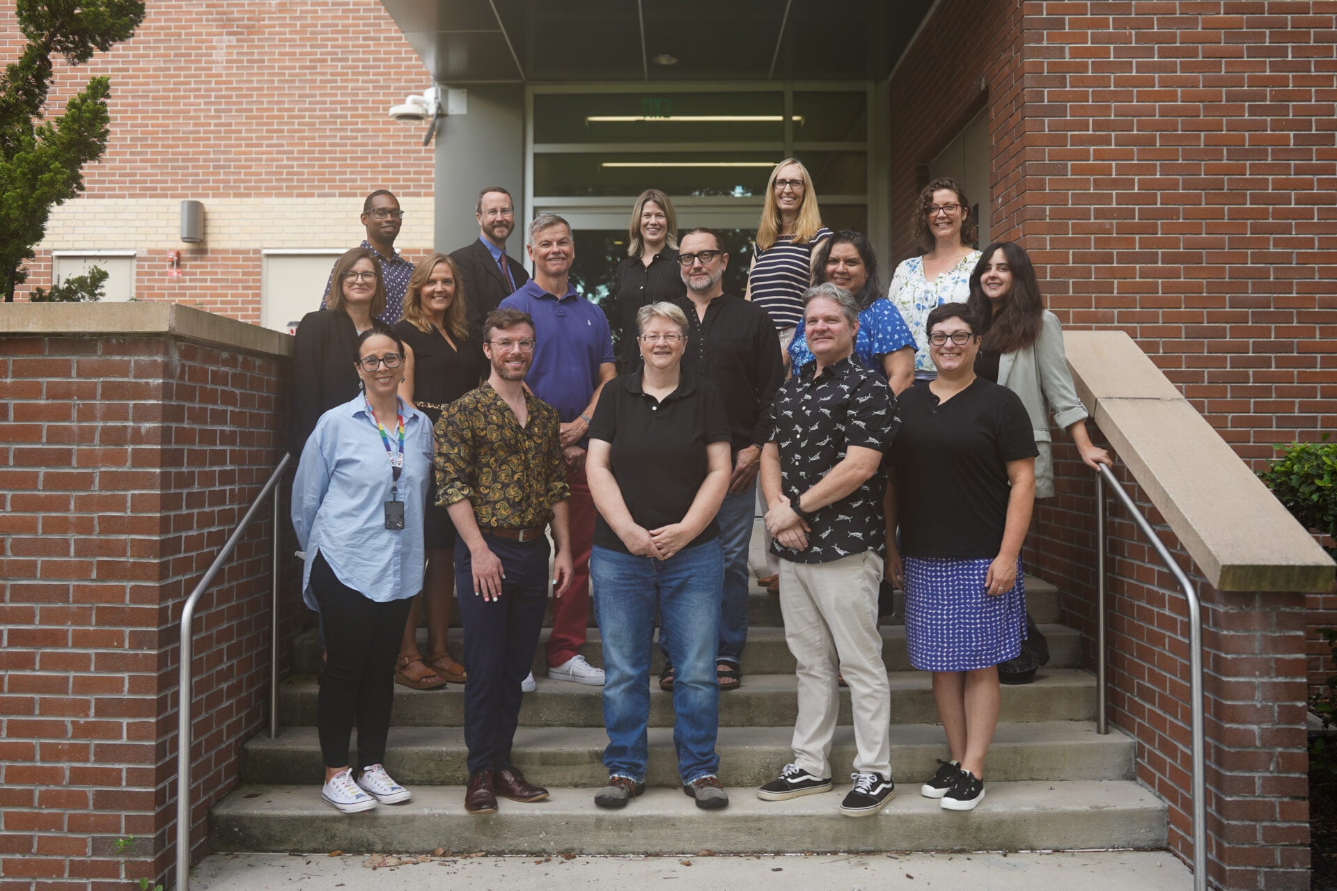 A group of sixteen adults stands on outdoor steps in front of a brick building, posing for a group photo.