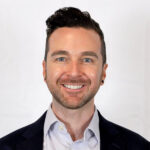 A man with short dark hair and a trimmed beard, wearing a dark blazer and light dress shirt, smiles in front of a plain white background.