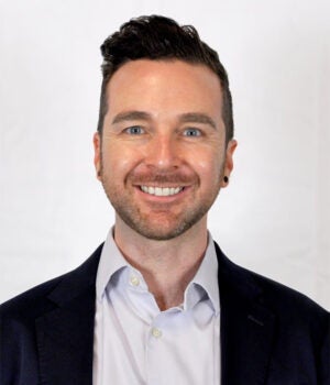 A man with short dark hair and a trimmed beard, wearing a dark blazer and light dress shirt, smiles in front of a plain white background.