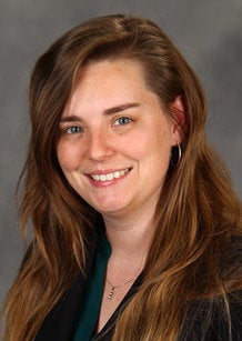 A woman with long brown hair, wearing a dark blazer and teal top, smiles at the camera against a gray background.