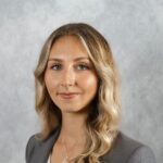 A woman with long, wavy blonde hair wearing a gray blazer and white top poses against a light gray background.