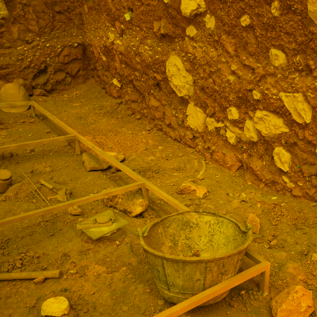 Excavation site with a metal bucket, tools, rocks, and a helmet on dusty ground against a rough earthen wall.