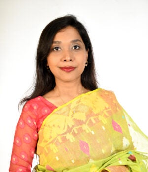 A woman with long dark hair wearing a patterned green and yellow saree with a red blouse stands against a plain white background, looking at the camera with a neutral expression.