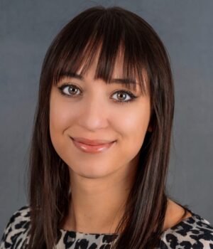 Woman with straight brown hair and bangs wearing a short-sleeve leopard print dress, standing in front of a plain gray background, looking at the camera and smiling softly.