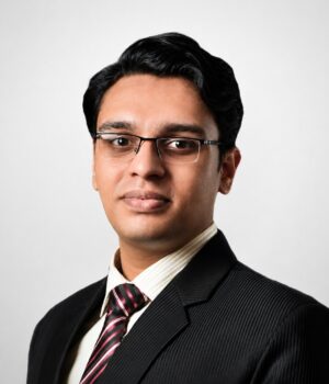 Man with short black hair and glasses wearing a dark suit, striped tie, and light shirt, posing for a professional headshot against a plain light background.
